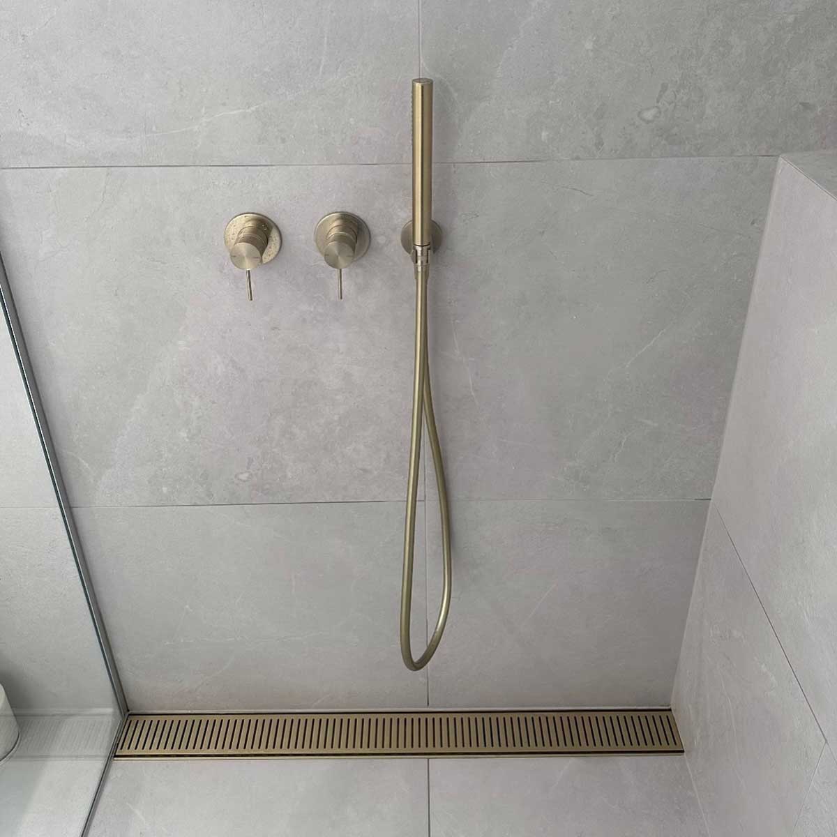 Shower setup with gold fixtures on a tiled wall and Lauxes Grates Custom Linear Shower Drain NeXT Generation - Matte Gold Installed against the tile wall and floor
