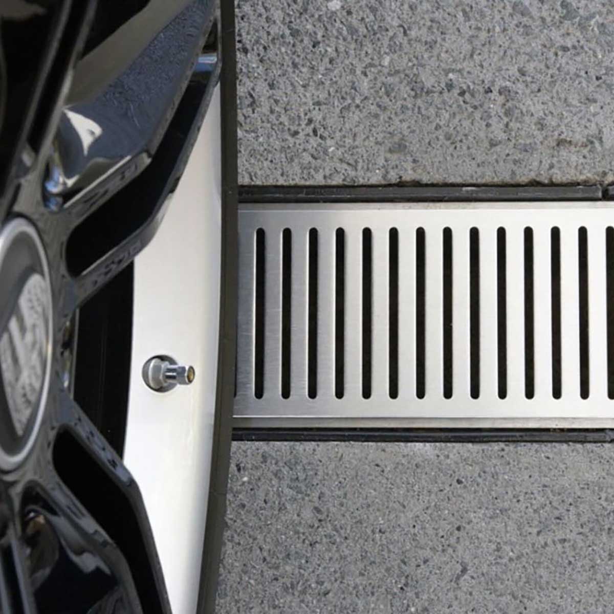 Close-up of a car tire over Lauxes Grates Custom Linear shower Drain NeXT Generation - Silk Silver on a concrete surface