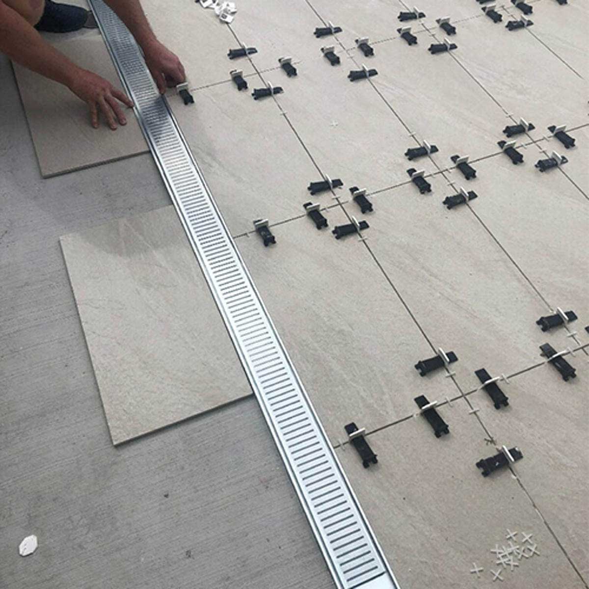 Close-up of a Person installing a Lauxes Grates Custom Linear shower Drain NeXT Generation - Silk Silver on a tiled floor with tools scattered around.