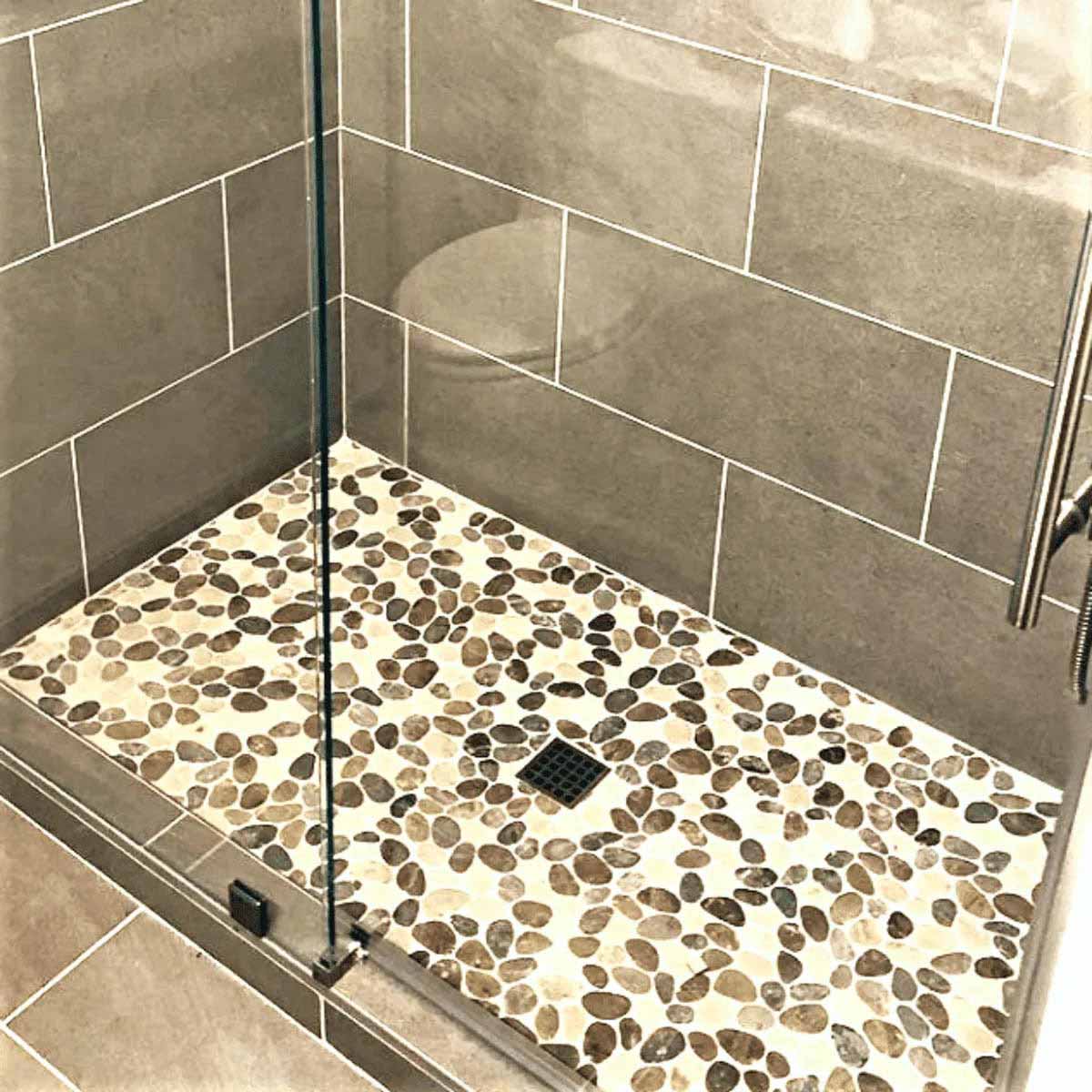 Modern bathroom with glass shower enclosure, pebble-patterned floor and tiled walls with Ebbe Drains Unique Square Shower Drain Grate - Frames