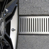 Close-up of a car tire over Lauxes Grates Custom Linear shower Drain NeXT Generation - Silk Silver on a concrete surface